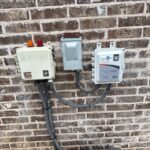 down-net_http20260611-122-tj7bca | Cyclone Septic & Plumbing Outdoor wall with three utility enclosures mounted on brick: left box has orange and red indicator lights, center small gray box, right Cyclone Septics controller with SeptiLink label and remote monitoring cables