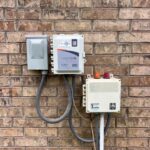 down-net_http20260611-131-q406mt | Cyclone Septic & Plumbing Outdoor electrical equipment mounted on a brick wall: a gray weatherproof box beside a white Septilink/Cyclone monitoring unit with cables and conduits nearby..
