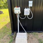 down-net_http20260611-202-rub8cy | Cyclone Septic & Plumbing Outdoor utility panels mounted on a dark metal wall, including a NuWater box and a SeptiLink/ Cyclone panel with cables and conduits, plus a concrete block at the base.