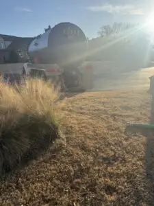 Large silver tanker truck parked on a quiet residential street with dry brown grass and sun glare in the foreground