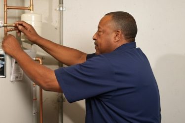 A man in a dark blue shirt is working on a plumbing system, adjusting copper pipes and a water heater, with his back partially visible.